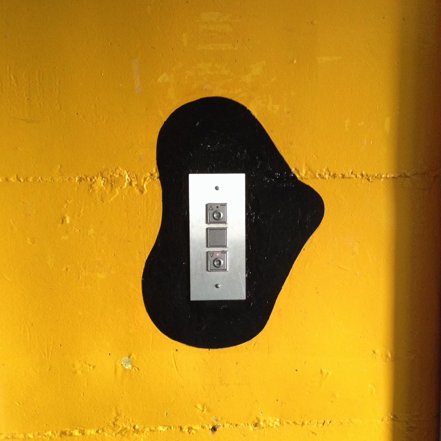 Les boutons d’appel de l’ascenseur sont cerclés d’une tâche noir qui se détache d’un mur jaune vif.